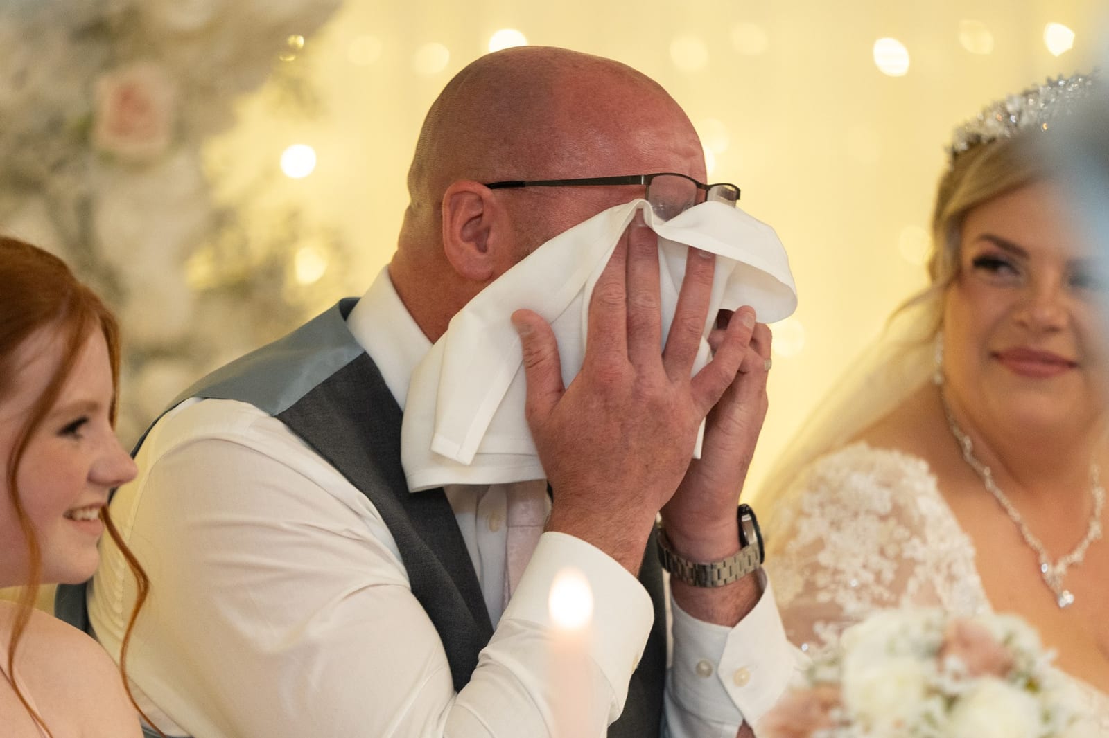 The groom wiping his eyes with a napkin during the speeches while the bride smiles beside him