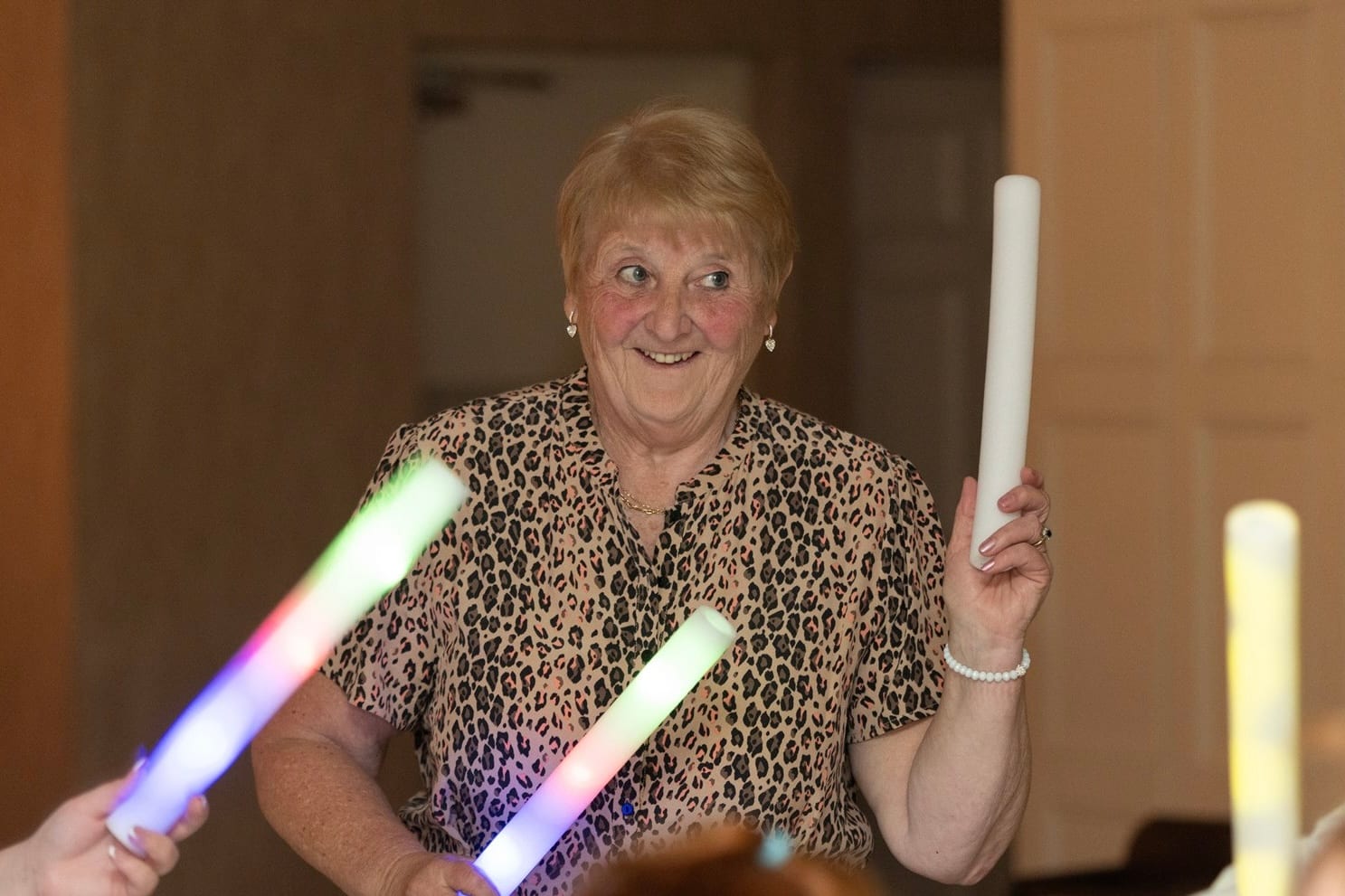 A grandmother in leopard print grinning on the dance floor, glow sticks in both hands