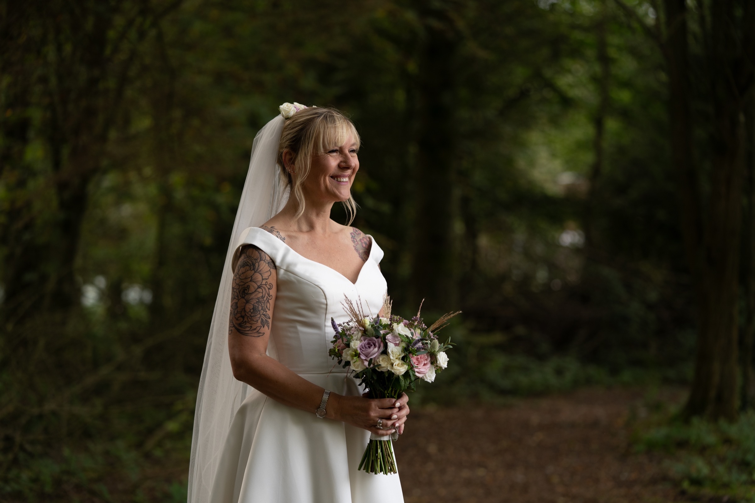 A bride stands in dappled woodland light, looking away — a quiet moment before the ceremony