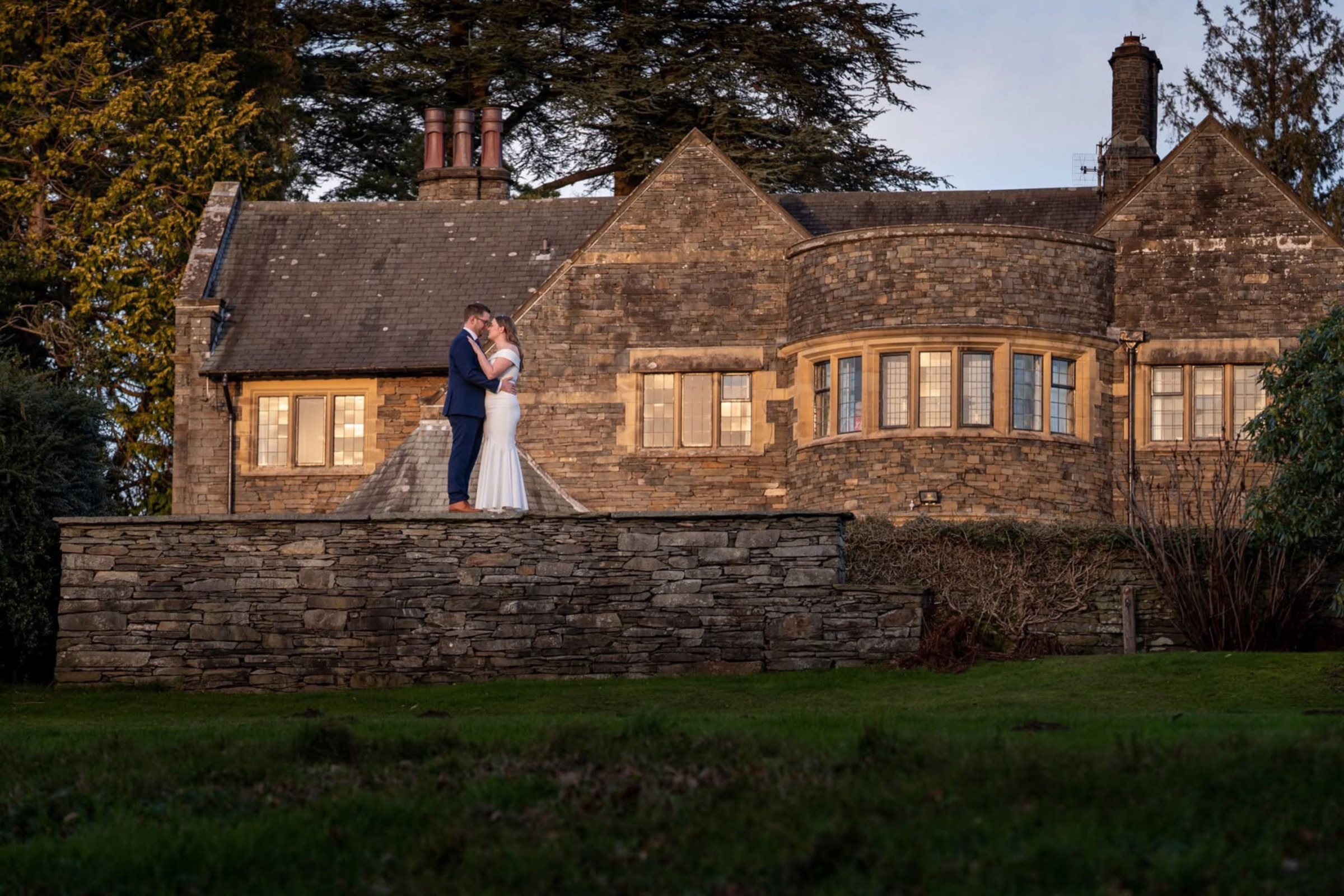 A couple stand close together in front of a floodlit stone manor house as evening falls