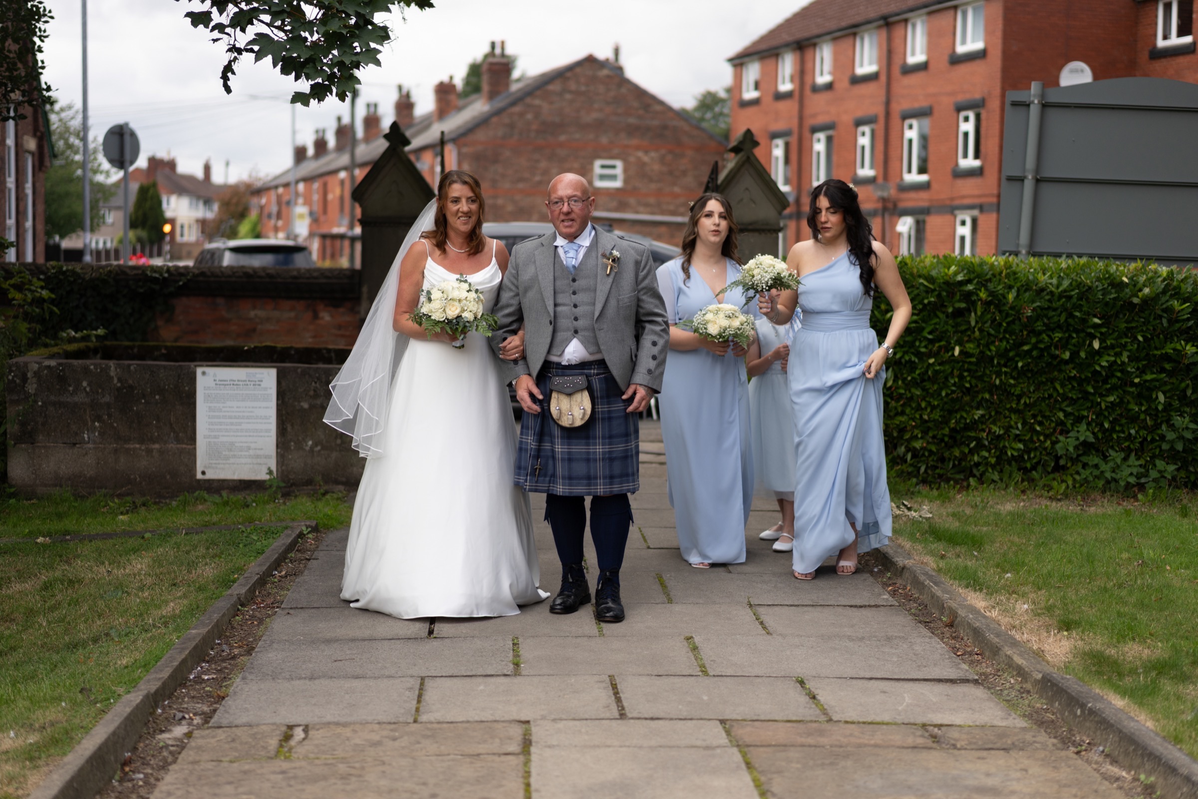 A bride walks to the church with her father in a kilt and two bridesmaids — composed, purposeful