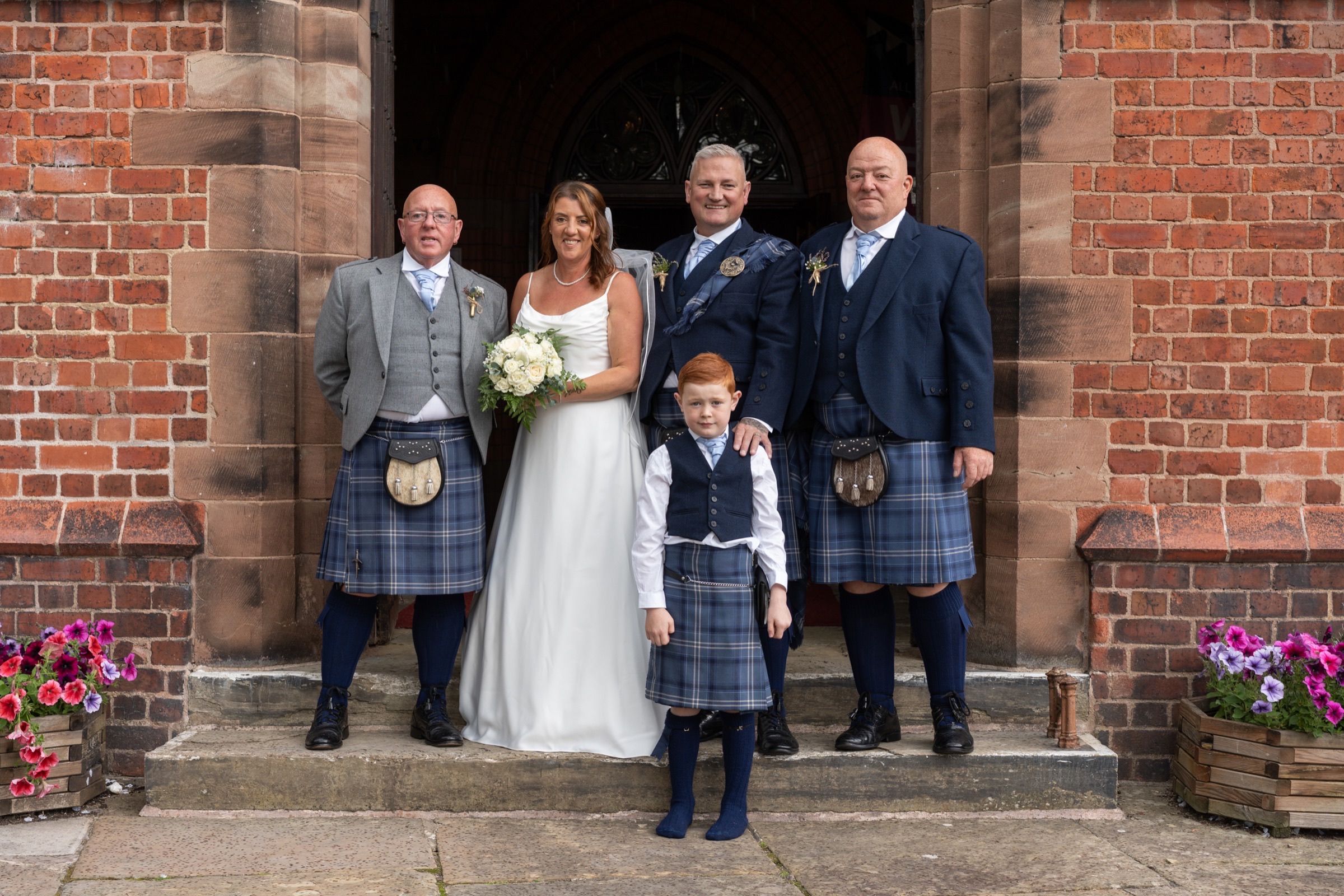 A wedding group — couple and family in kilts — standing together at the church entrance, relaxed and smiling